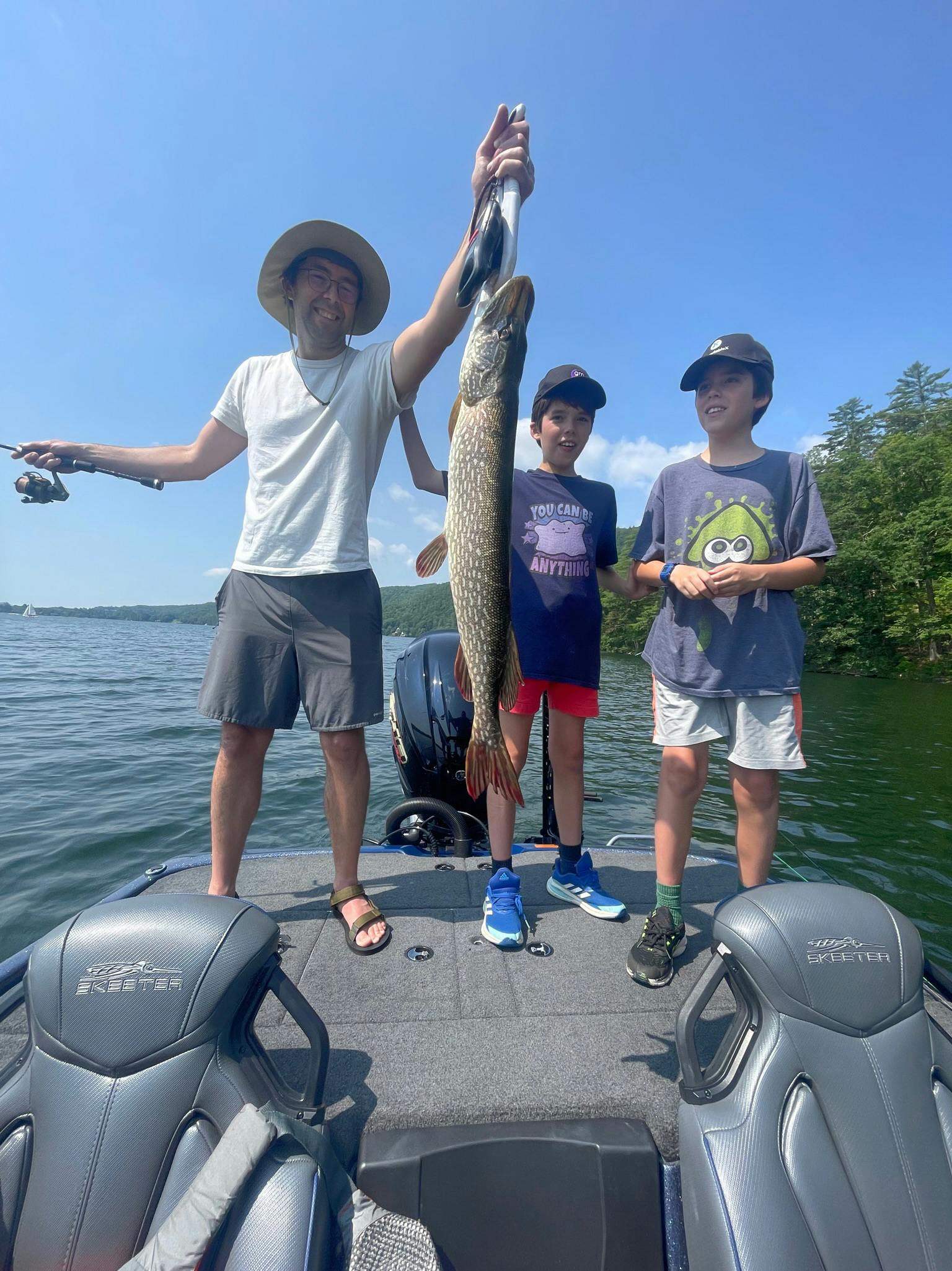 Father in white shirt, grey shorts, and tevas holds northern pike with two twin boys each wearing navy t-shirts and black baseball caps on boat