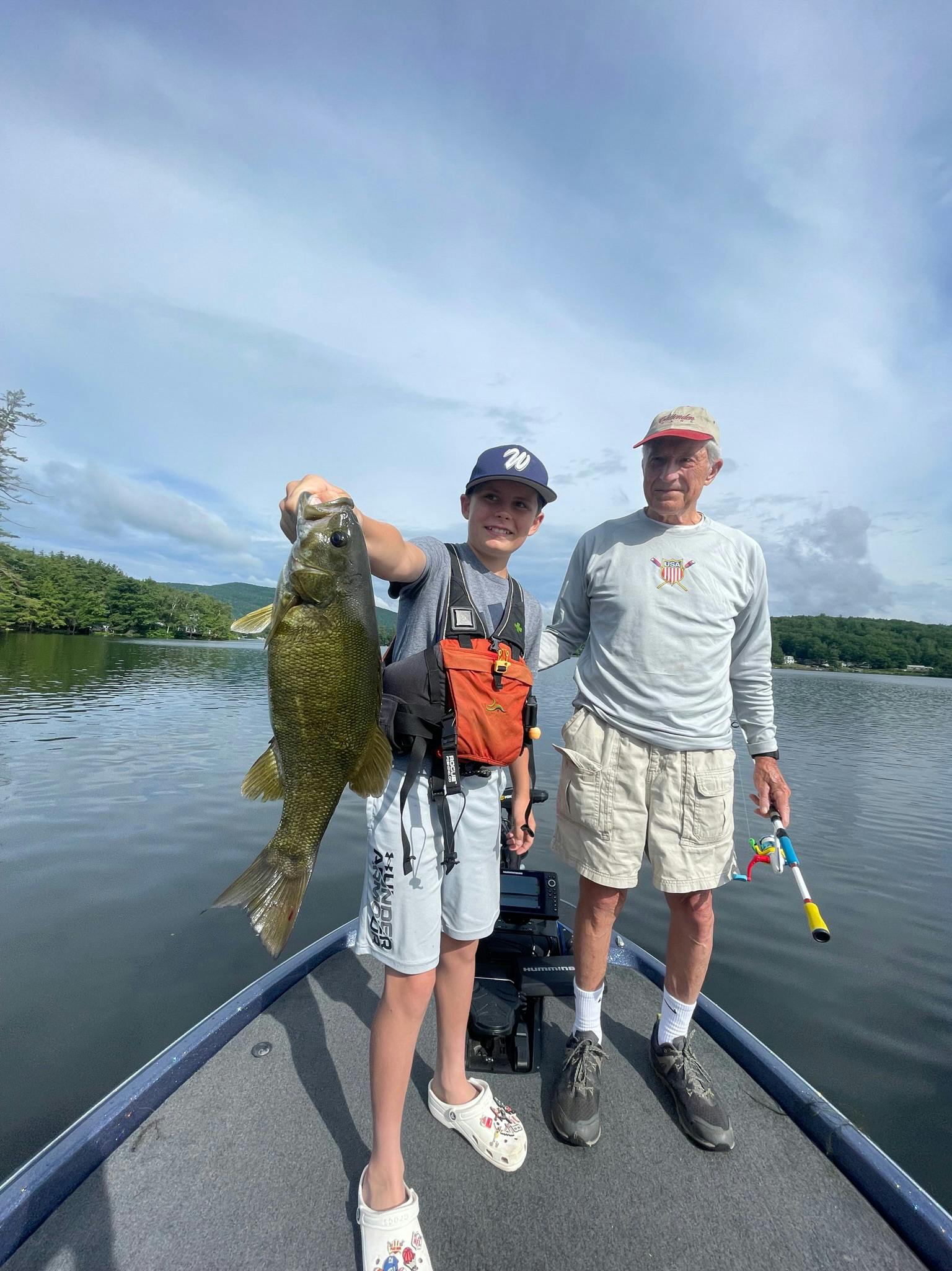 Boy in blue baseball hat and white crocs holding smallmouth bass next to grandfather wearing grey shirt and khaki cargo shorts on blue boat