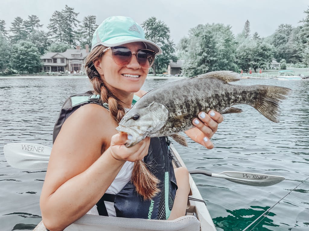 Woman in blue Adidas hat holding one fish while smiling in kayak