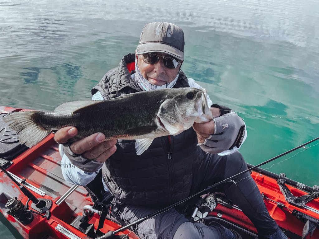 Man in white sweatshirt and grey Under Armour hat holding one fish while smiling in kayak