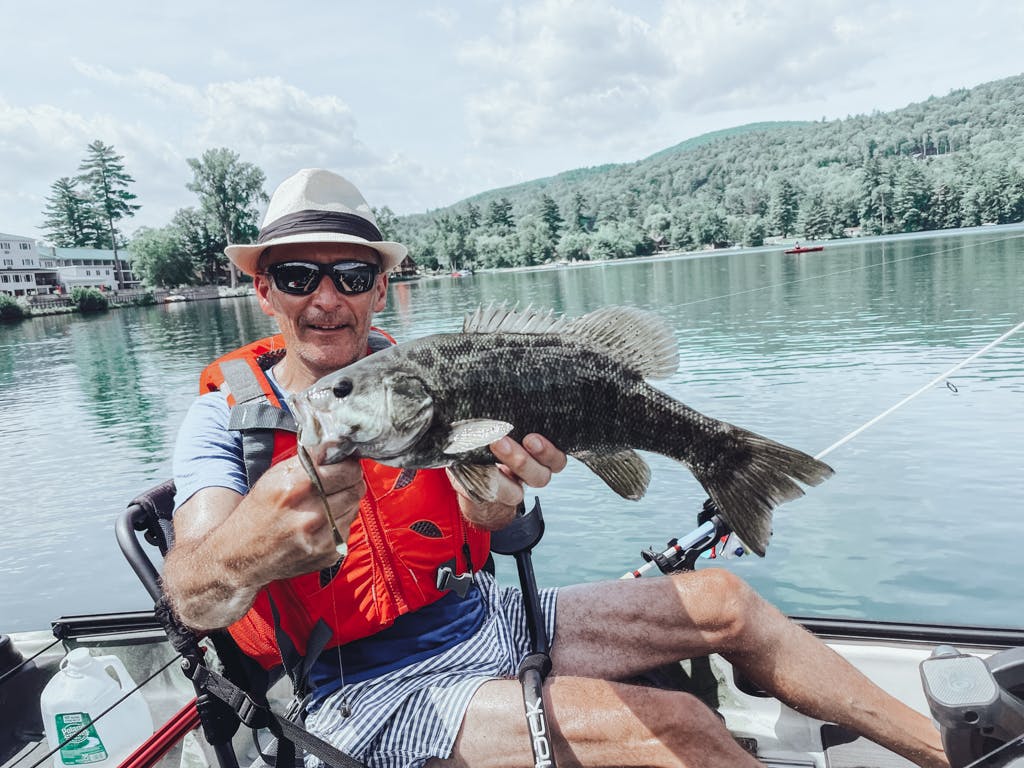 Man in blue T-shirt and fedora holding one fish while smiling in kayak