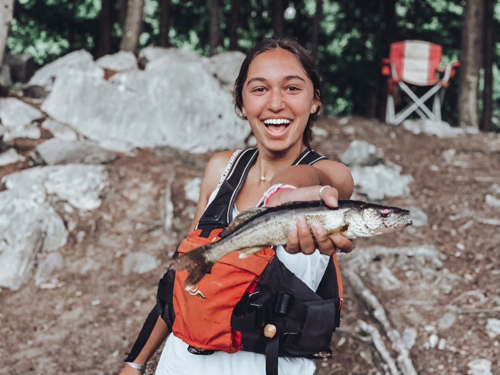 Young woman in white tank top holding one fish while smiling on lake shore