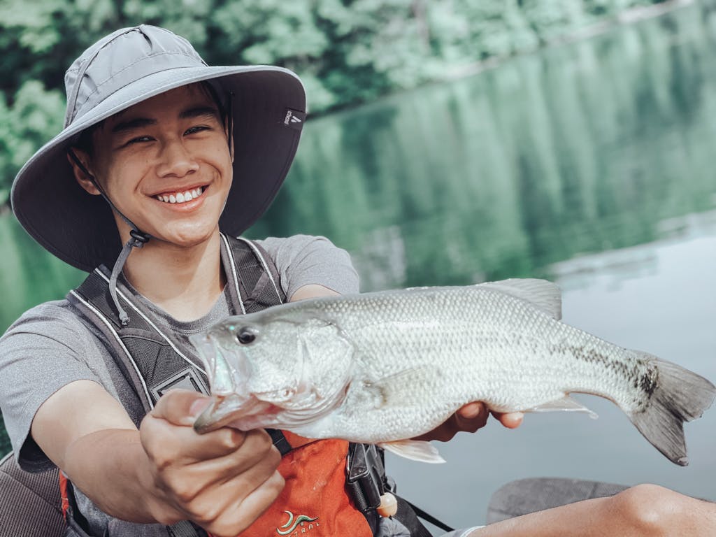 Young man in grey T-shirt and large sun hat holding one fish while smiling in kayak