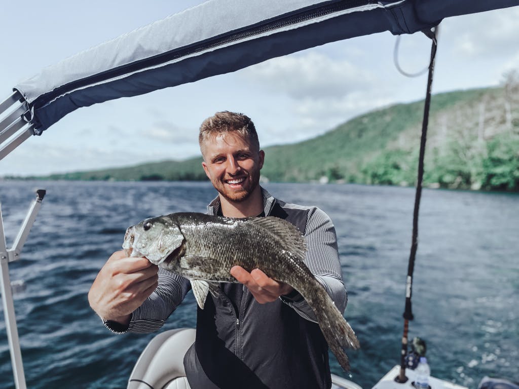 Man in grey and black quarter-zip a holding one fish while smiling on boat