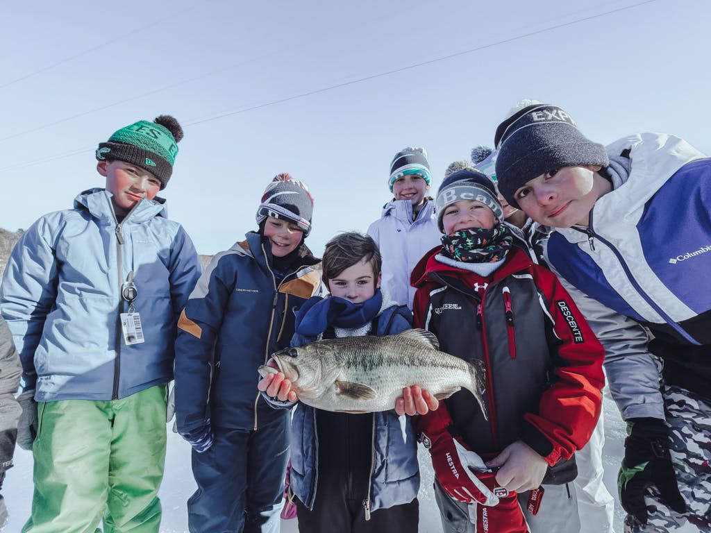 Boy #1 with a scarf holding a fish on a frozen lake surrounded by other boys while smiling