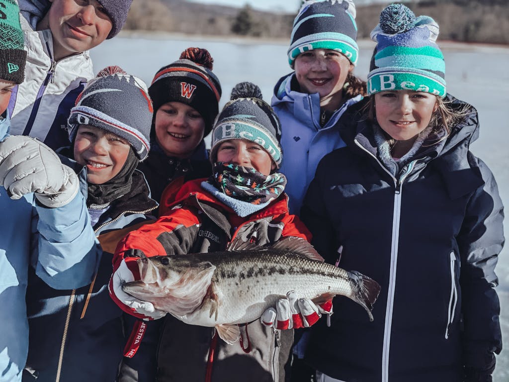 Boy #2 with a scarf and an LL Bean hat holding a fish on a frozen lake surrounded by other boys  while smiling