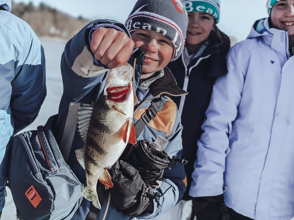 Boy #3 with a scarf and an LL Bean hat holding a fish on a frozen lake surrounded by other boys  while smiling