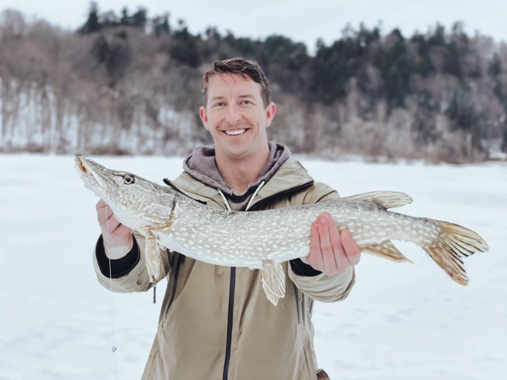 Middle-aged man in a beige jacket holding a fish on a frozen lake while smiling