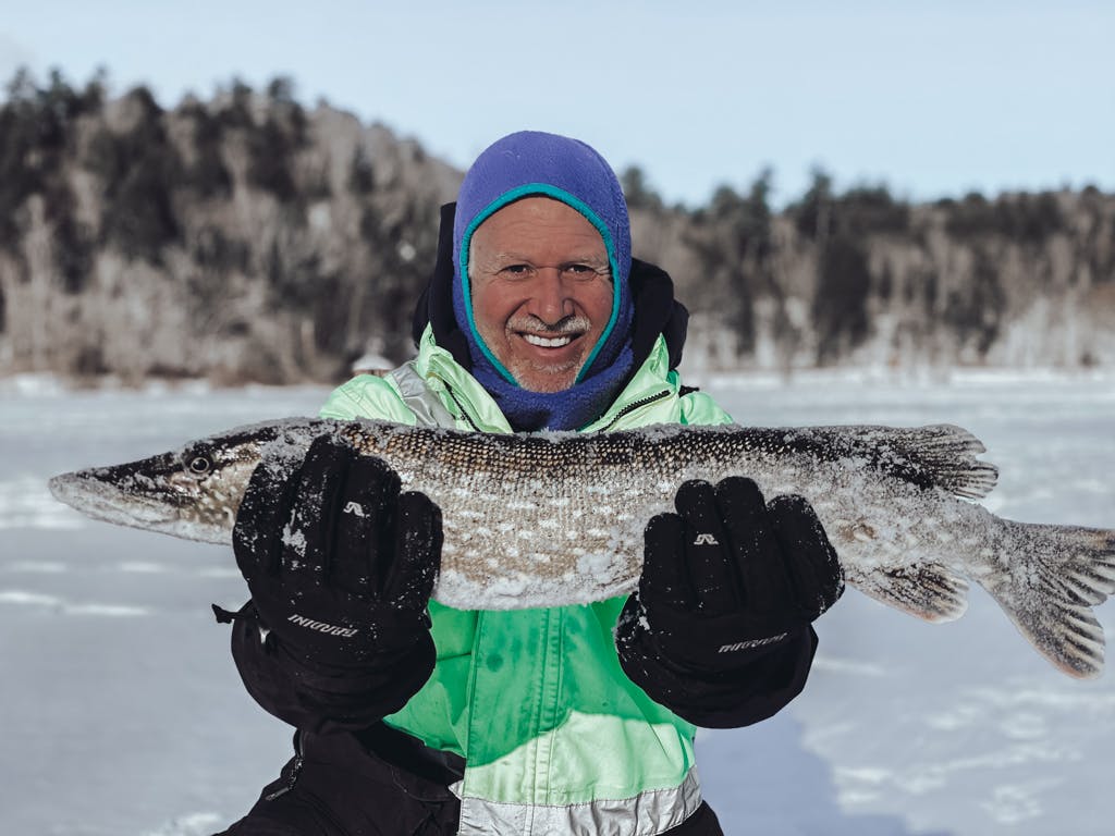 Older man in a green jacket and blue balaclava holding a fish on a frozen lake while smiling