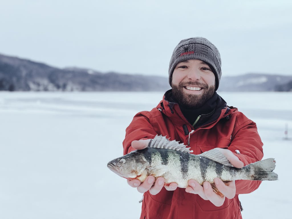 Middle-aged man in a red jacket and grey hat holding a fish on a frozen lake while smiling