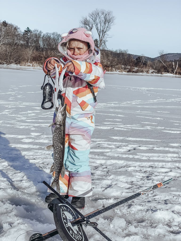 Young girl in a warm jacket and pink sweatshirt holding a fish on a frozen lake while smiling