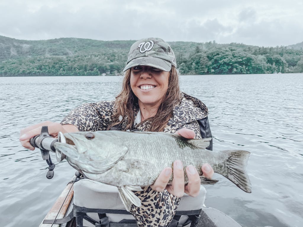 Young woman in Washington Nationals hat and cheetah-print jacket holding one fish while smiling in kayak