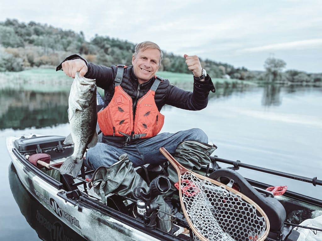 Middle-aged man in a black jacket and jeans holding one fish in a kayak while smirking