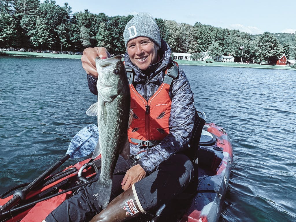Middle-aged woman in a grey jacket and green hat holding one fish while smiling in a kayak