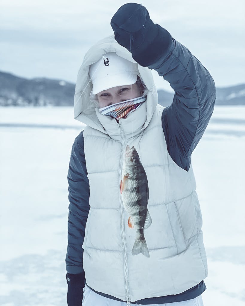 Woman wearing black jacket, white vest, white hat, and black gloves holding a fish on a frozen lake