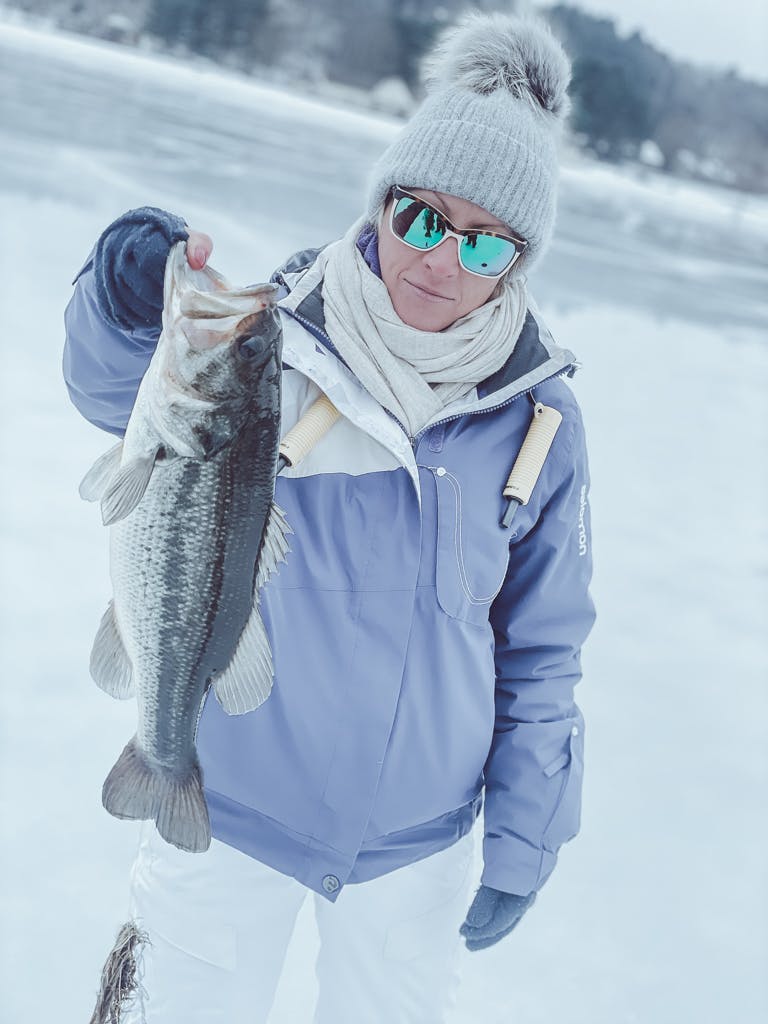 Woman in a purple solomon jacket, white scarf, grey beanie, and sunglasses holding one fish while smirking