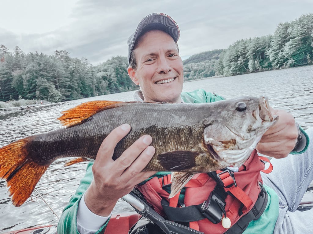 Man in green shirt and american hat holding one fish while smiling in kayak