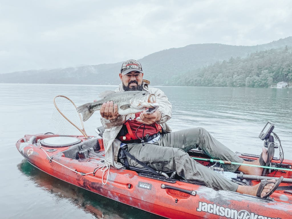 Man in grey jacket and flip flops holding one fish while smiling in kayak